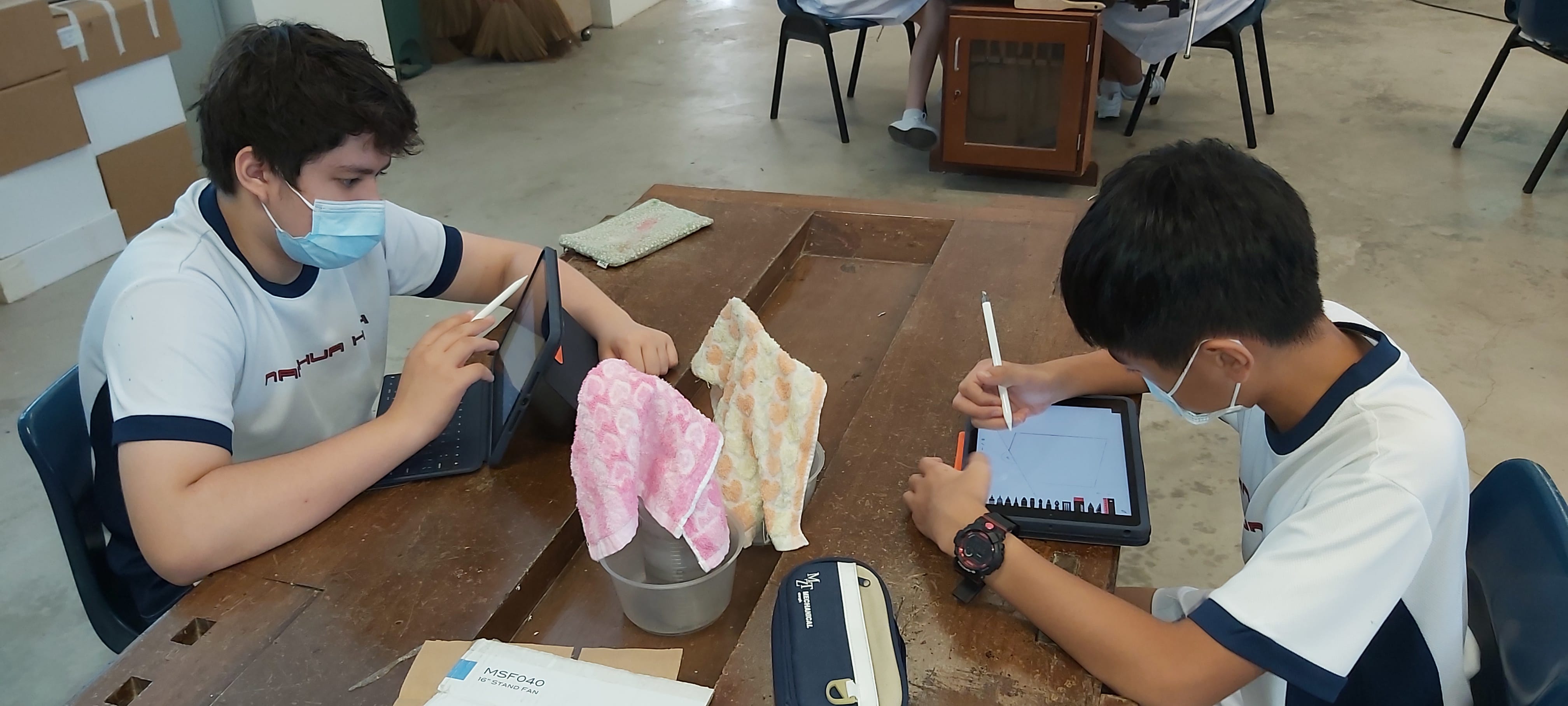 Two masked students sit at a wooden table using tablets, with cloths and a bucket between them.